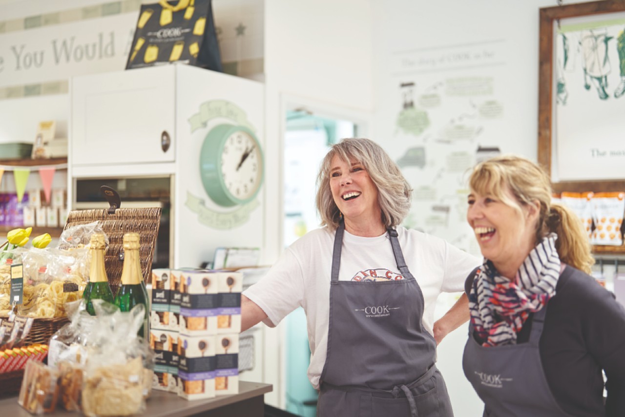 Two people behind a shop counter with food products on display